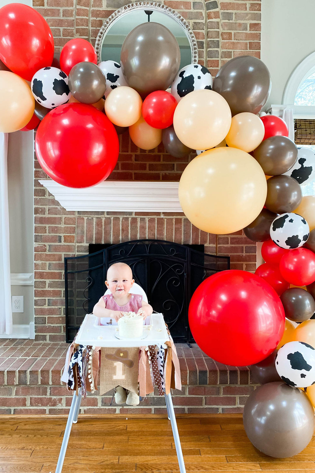 Cute little baby boy in overalls celebrating his first birthday with a first rodeo themed party. Sitting under a balloon garland with his smash cake in his high chair ready to celebrate